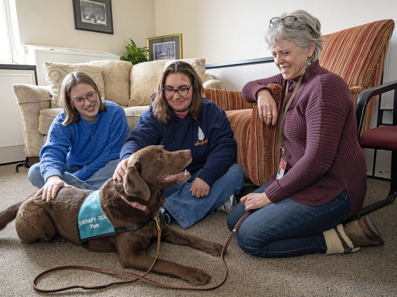 A person kneels on the floor next to two other people sitting cross-legged and petting a therapy dog with a vest and a leash on. Behind the group are two chairs and a couch.