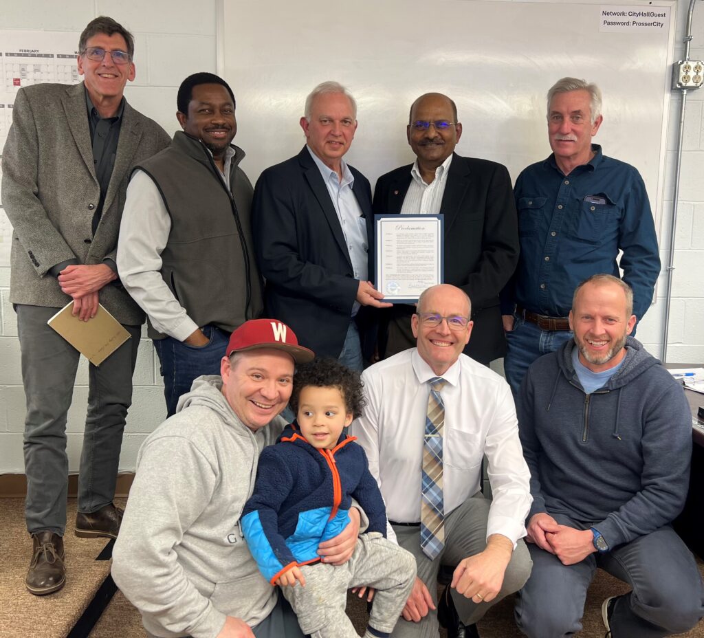 A group of five people stands. Three more people squat in front of them, one holding a child. Two people who are standing hold a framed proclamation.