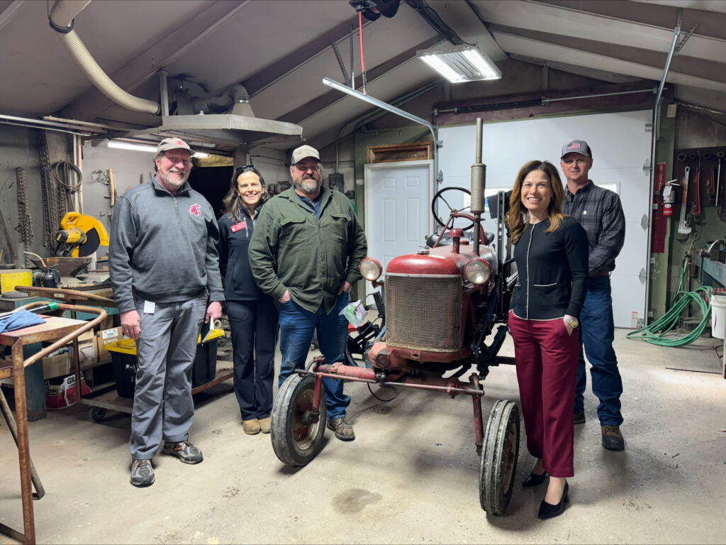 Five people stand near an antique tractor inside of a garage. 