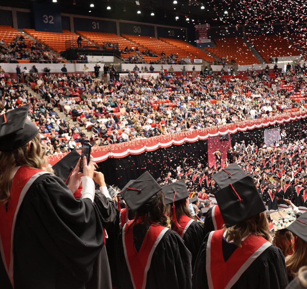 The back of graduates standing in a crowded Beasley Coliseum as confetti rains down at graduation.
