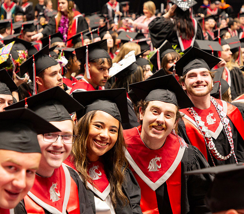 A group of students sitting, some smiling at the camera, while wearing caps and gowns at graduation.
