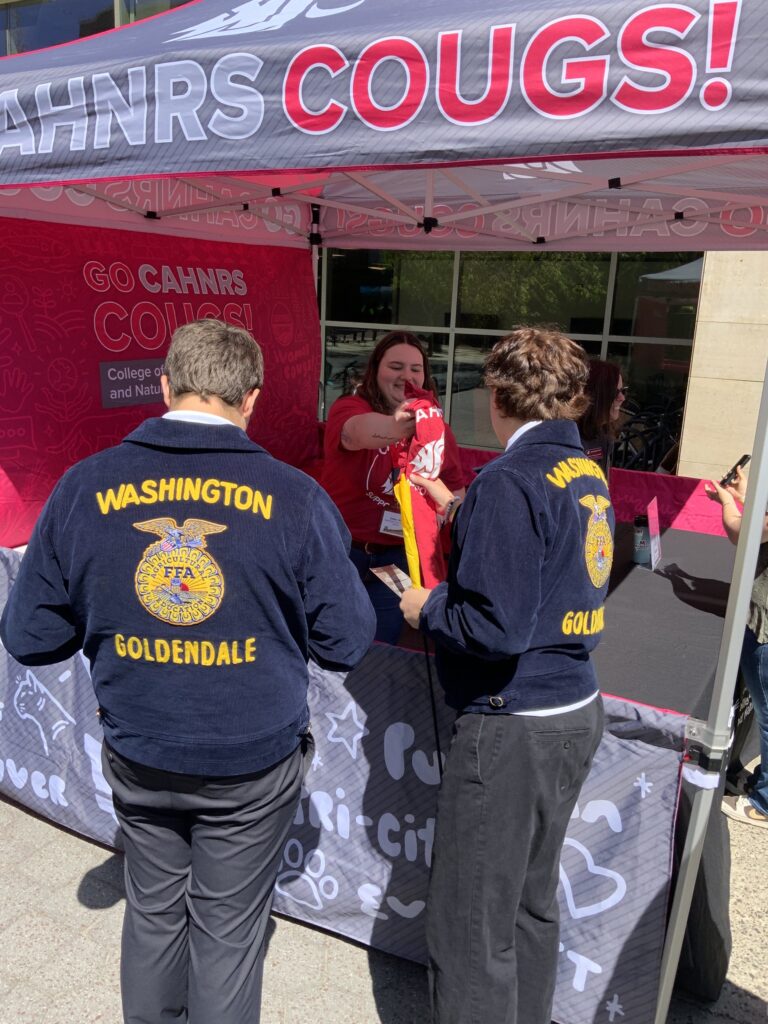 A person stands in a WSU CAHNRS-themed tent and hands out t-shirts to two FFA students.