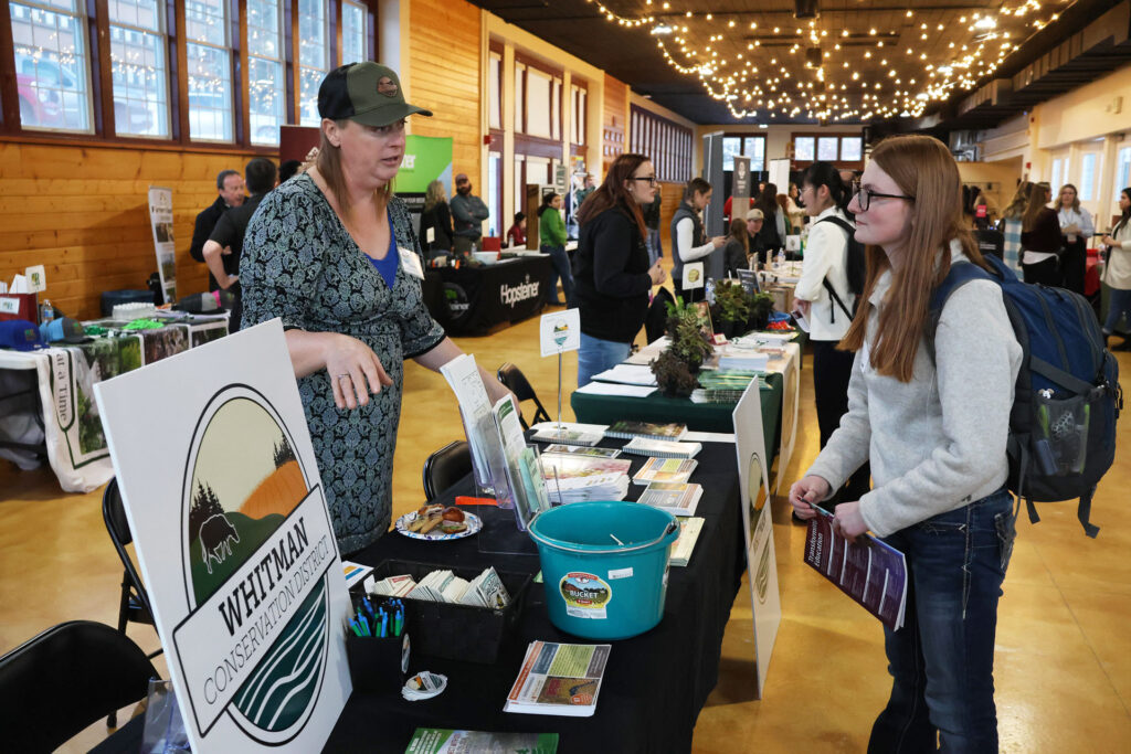 Two people talk while standing with a table between them. The table is covered with papers, fliers, stickers, a blue bucket, a sign that says Whitman Conservation District, and more. Other people stand and talk to each other near tables in the background. 