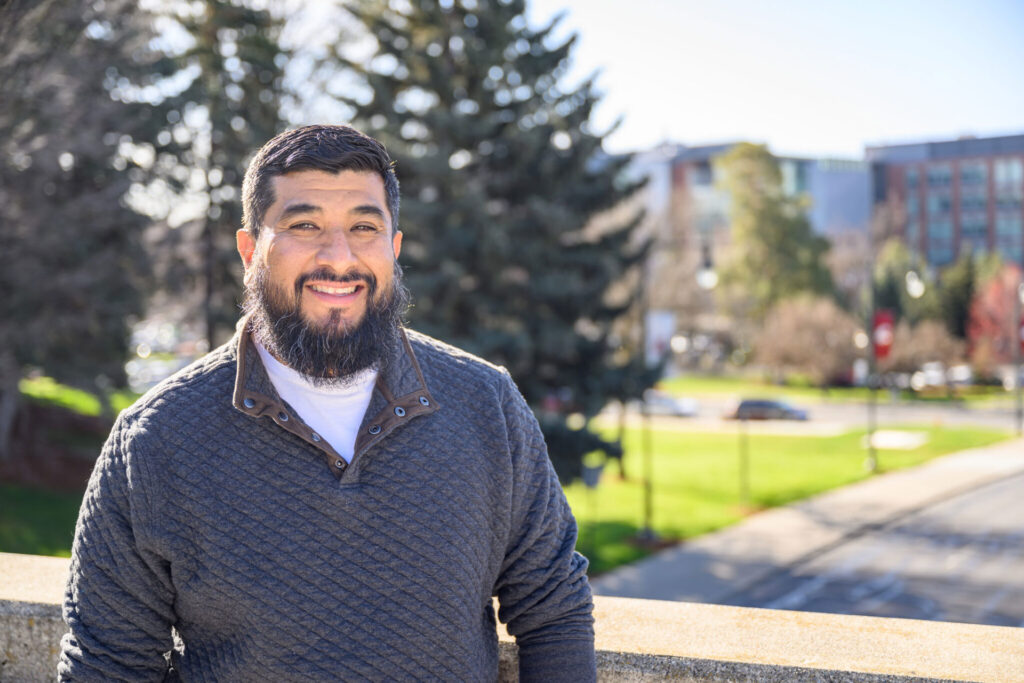 Professional head shot of Junior Gomez standing outside. Trees are in the background behind him