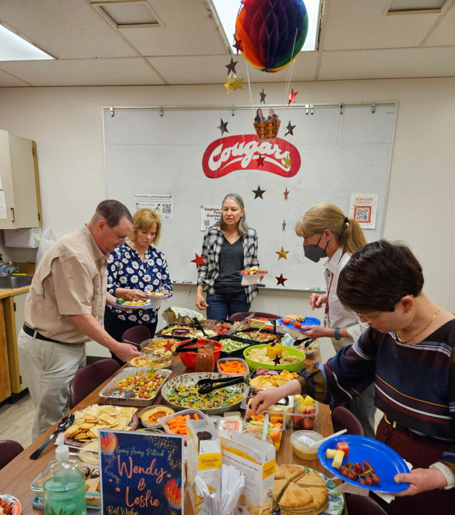 Five people fill their plates while standing next to a table of food at a potluck.