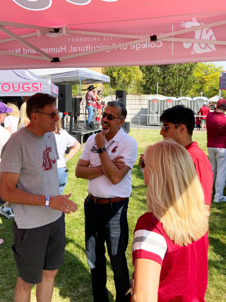 Four people stand talking under a tailgate tent with a band playing behind them.