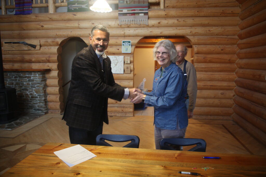Two people shake hands while smiling and standing next to a table with a paper and pens on it. They are inside of a log cabin. Behind the two people is another person, barely visible.