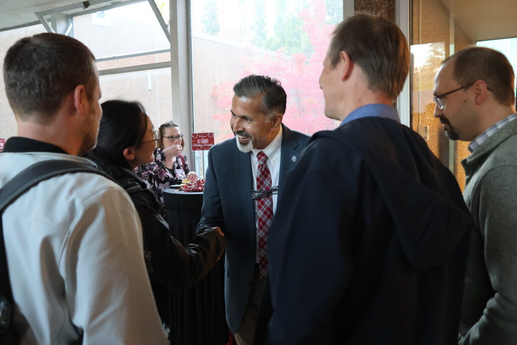 At a reception, Khosla, center, smiles and greets several colleagues; woman is standing at a table in the background.