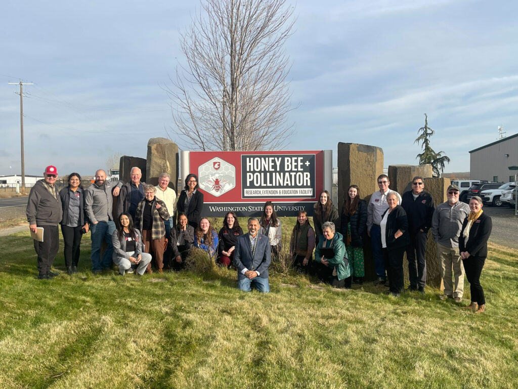 22 people stand outside on grass next to a sign that reads "Honey Bee + Pollinator Research, Extension, and Education Facility."