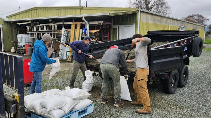 Four people working to fill sandbags with sand from the back of a trailer. Next to them is a buliding with ladders and other pieces of equipment.