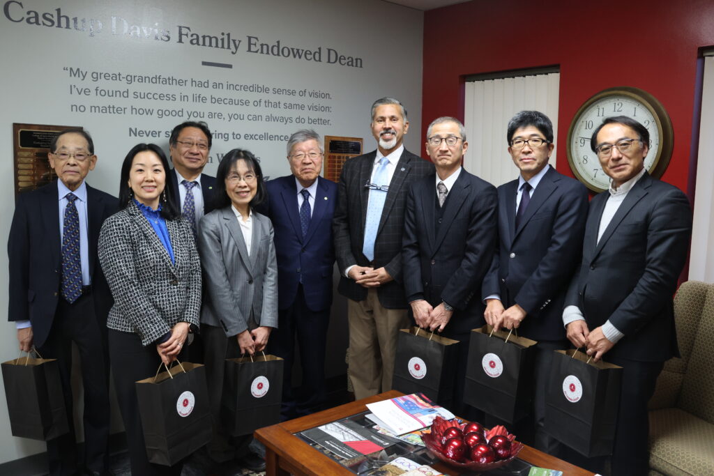 A group of nine people stands together, some holding paper gift bags. On the wall behind them, text reads "Cashup Davis Family Endowed Dean" with a quote underneath.