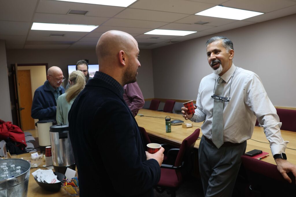Two people hold paper cups while speaking to one another in a conference room. A long table is behind them. Nearby, a large metal container of coffee sits on a table alongside other cups, bottles of water, hot chocolate packets, and tea packets. A few other people stand in the background.