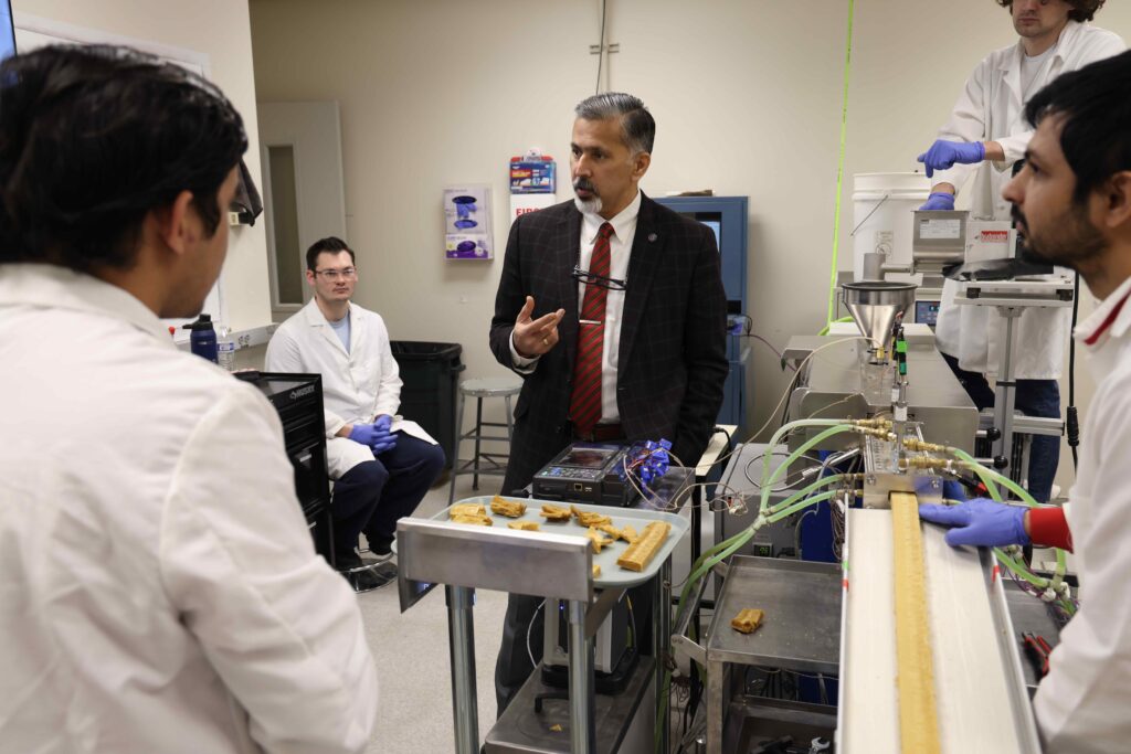 A person gestures while standing in a food science lab. Several people in lab coats and wearing latex gloves stand nearby. There is food processing equipment and a tray full of food samples.