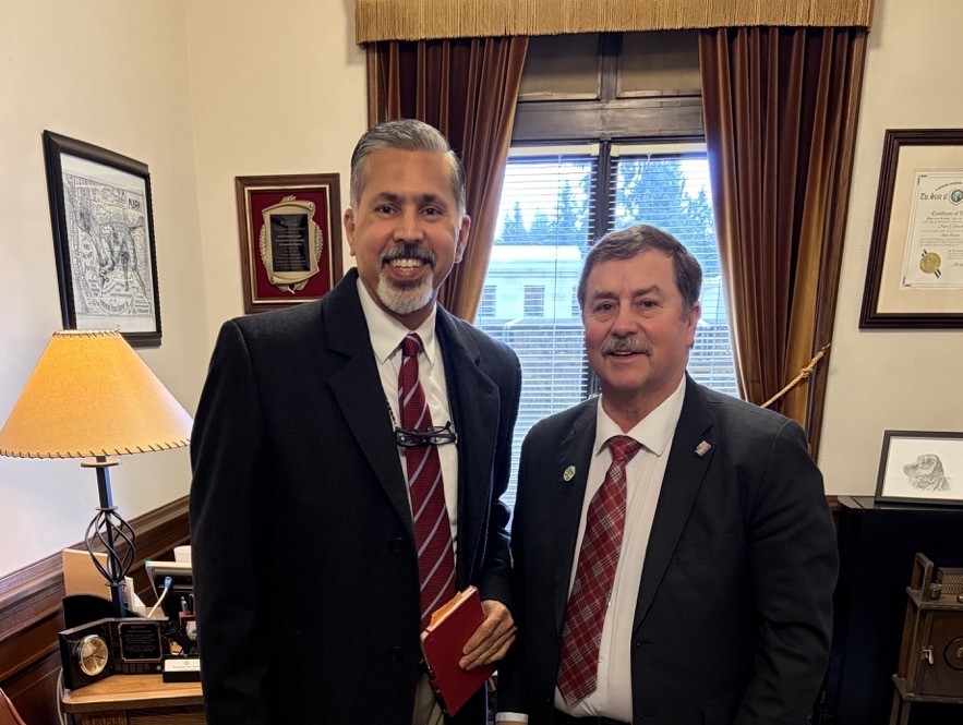 Raj Khosla and Mark Schoesler stand together smiling while wearing suits and ties. Raj is holding a notebook.