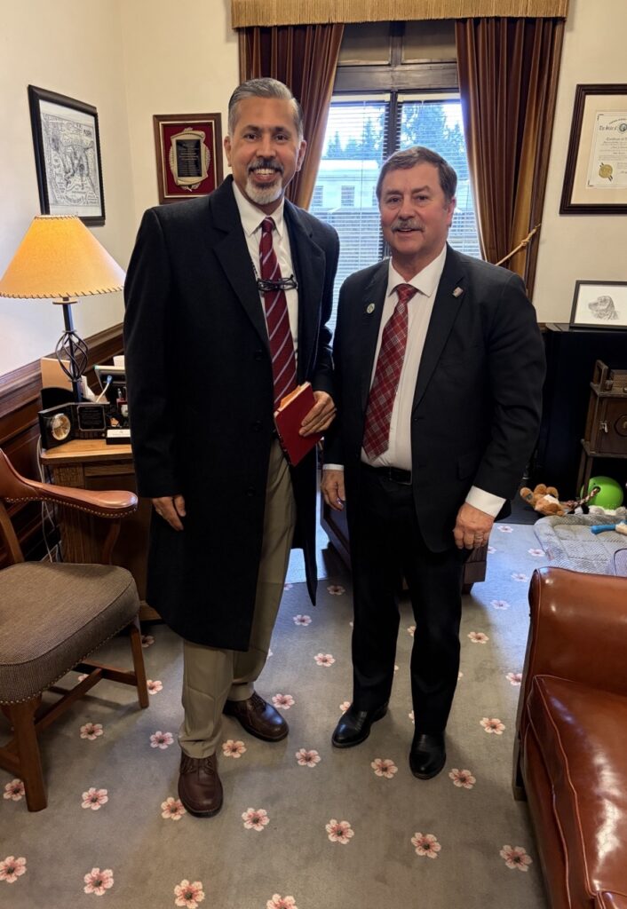 Raj Khosla and Mark Schoesler stand together smiling while wearing suits and ties. Raj is holding a notebook.