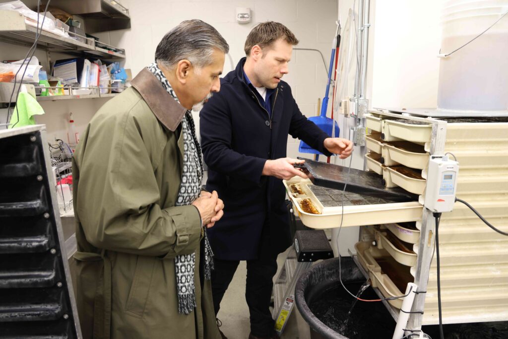 Two men stand together in a lab setting as one pulls a tray out of a stack of trays.
