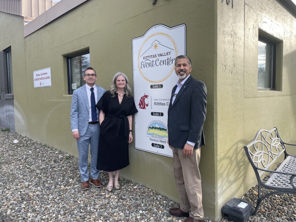 Three people stand next to a sign on the outside of a building. The sign reads "Kittitas Valley Events Center, Suite 1, WSU Extension Kittitas County, Suite 2, Kittitas County Noxious Weed, Suite 3."