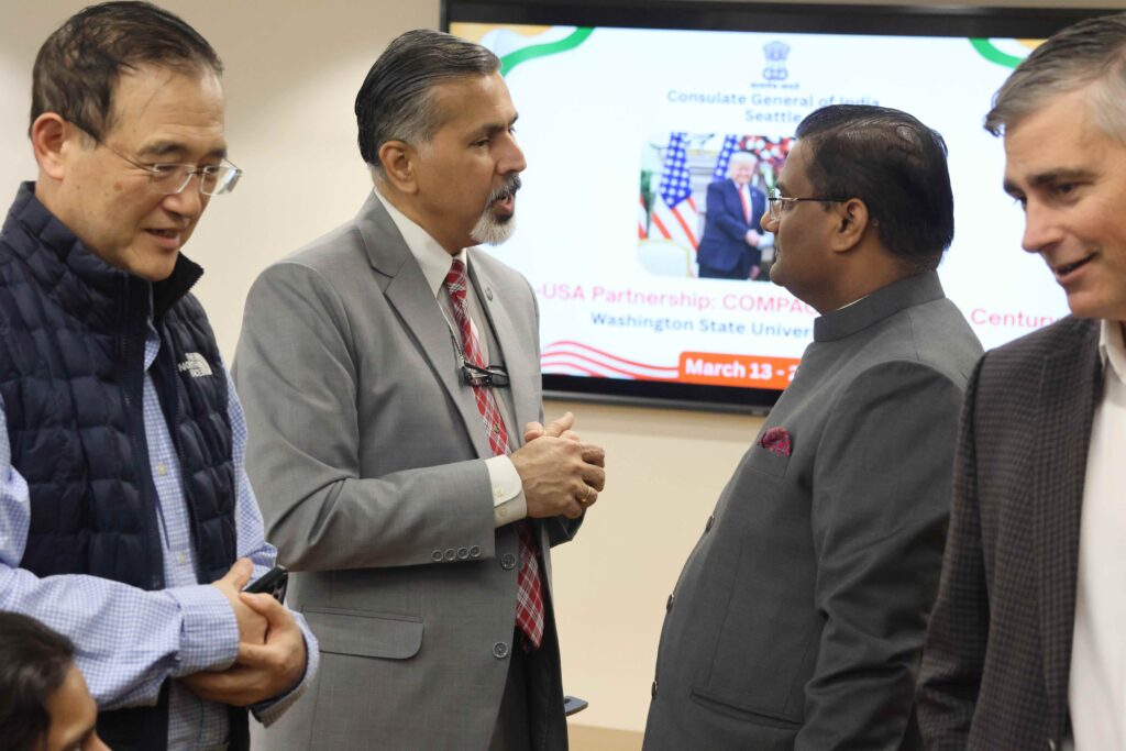 Raj Khosla and Prakash Gupta stand together, speaking. A few others are standing in front of them. In the background is a slide projector displaying a slide with information about the Consulate General of India.