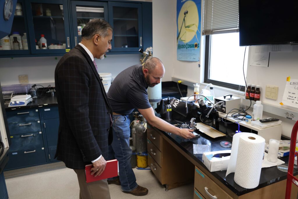 A person leans down to touch a microscope on a table in a lab setting. Raj Khosla stands behind him and looks down while holding a notebook.