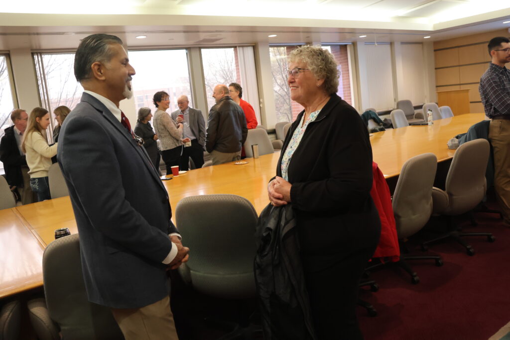 Raj Khosla speaks to Sandra Brown in a conference room with a large table and chairs. Several other people near windows in the background.