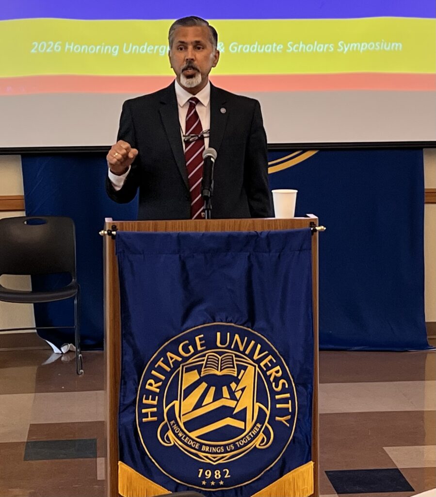 Raj Khosla stands at a podium, gesturing while he speaks. A banner on the podium reads Heritage University.
