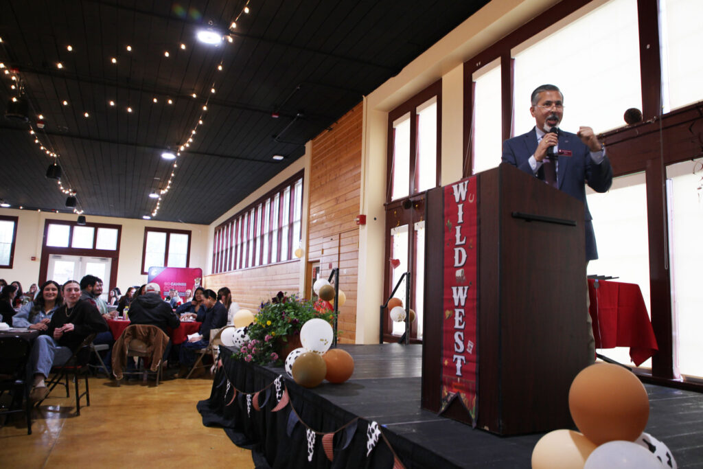 Dean Khosla gestures while speaking at a podium on a stage during the CAHNRS Student Awards Ceremony. Several people seated at round tables are looking at him. On the podium is a sign that reads Wild West. There are balloons and other decorations on the stage.