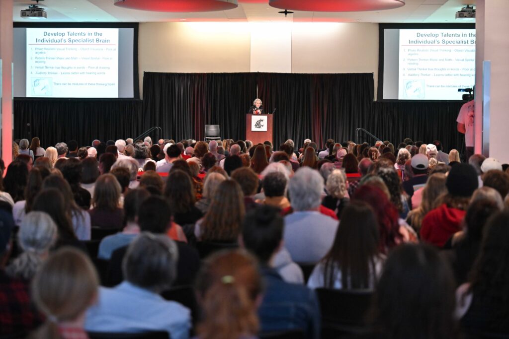The back of a large, seated audience listening to Temple Grandin, who stands at the front of the room on stage at a podium. The podium has a WSU cougar head logo on it. Two projector screens are pictured near the stage. The title on the projector screens reads "Develop talents in the individual's specialist brain"
