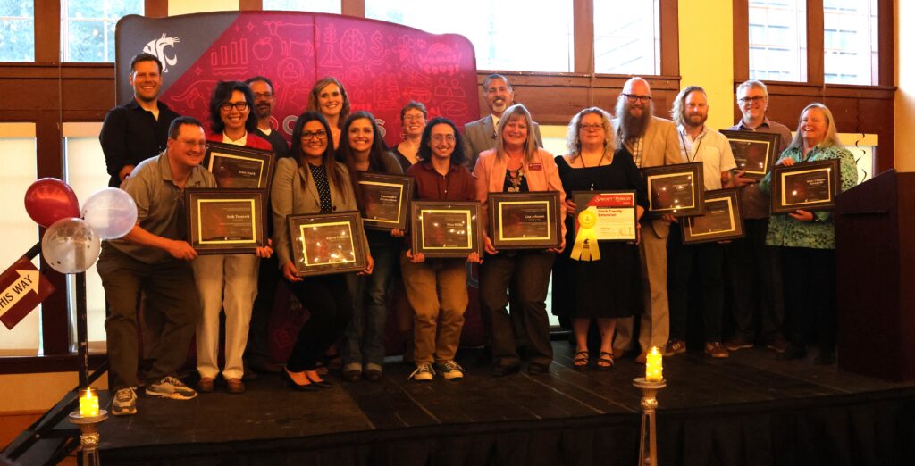 A large group stands on a stage in front of a WSU CAHNRS-themed background and windows. Most of the people in the group are holding framed awards.