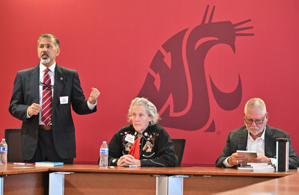Dean Raj Khosla, Temple Grandin, and Gordon Davis at a table in front of a background with the WSU cougar head logo. Raj stands and gestures while speaking. Temple and Gordon sit at the table.