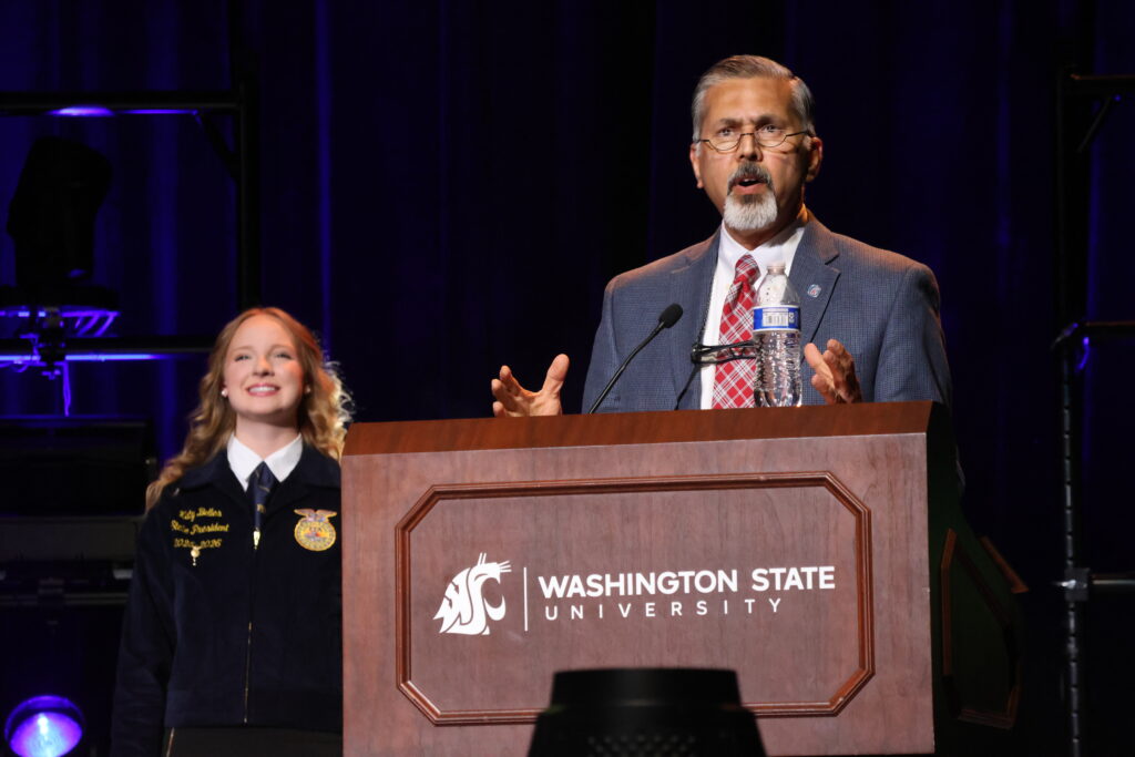 Raj Khosla speaks at a podium with the WSU cougar head logo and the words Washington State University on it. In the background, an FFA student stands, smiling.