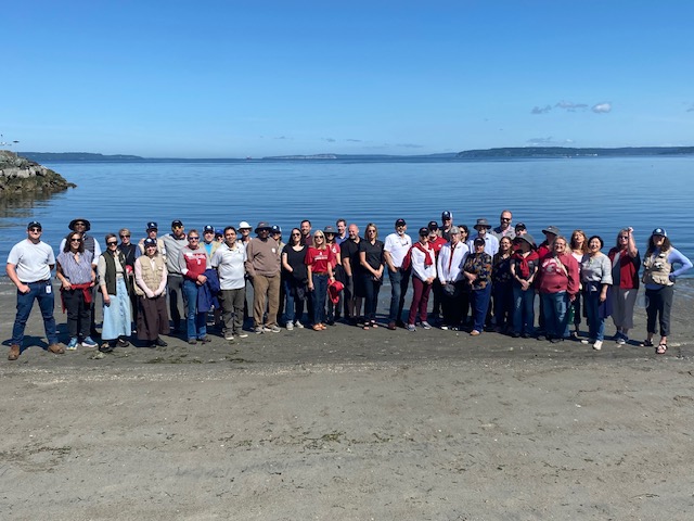A large group stands on a beach with water and blue skies behind them.