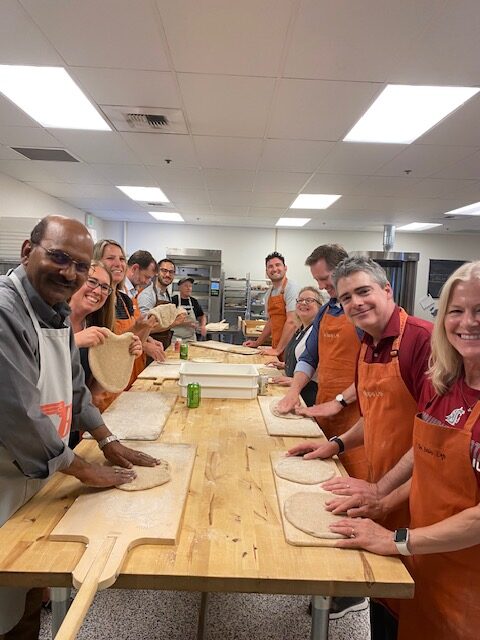 Several people on either side of a long wood countertop smile at the camera while working with pizza dough.
