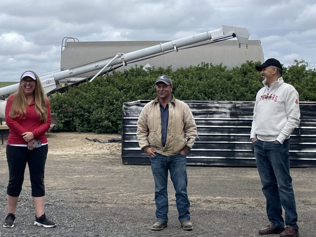 Three people stand together smiling. Behind them are trees and farm equipment.