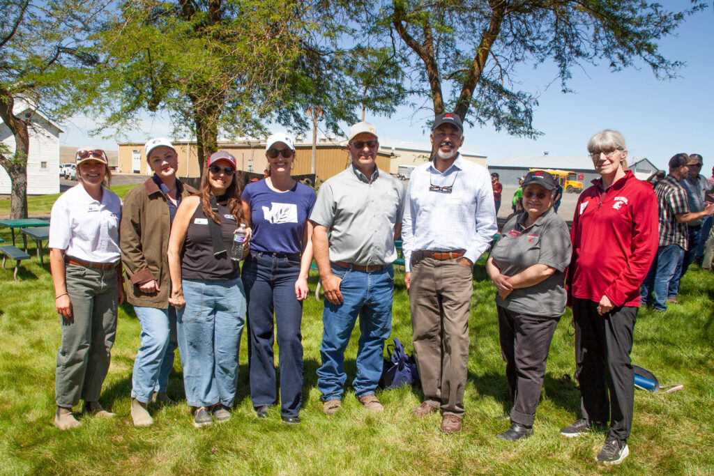A group of eight people stands together outside near trees, smiling.