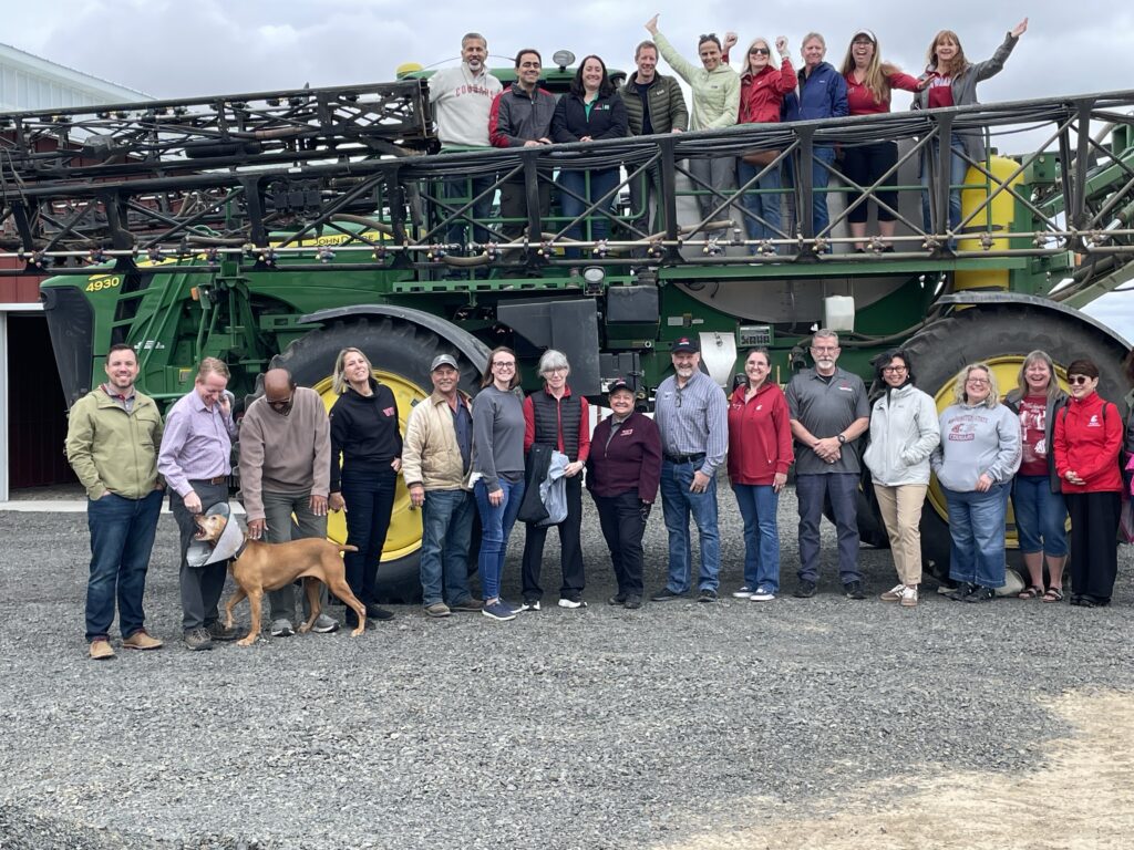 A large group of people and a dog stand on the ground in front of a large tractor. Several more people stand on top of the tractor.