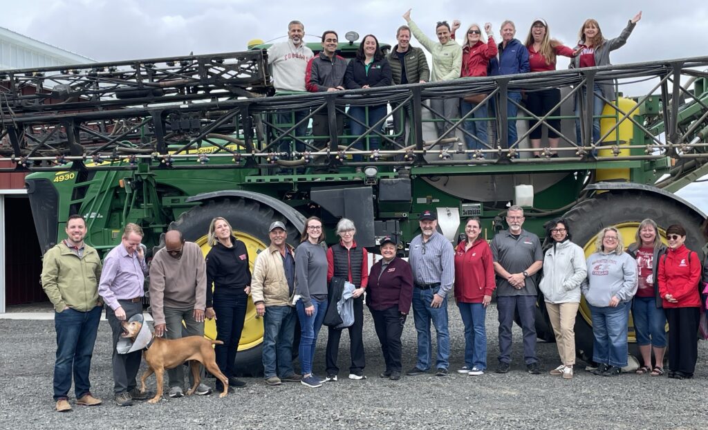 A large group of people and a dog stand on the ground in front of a large tractor. Several more people stand on top of the tractor.