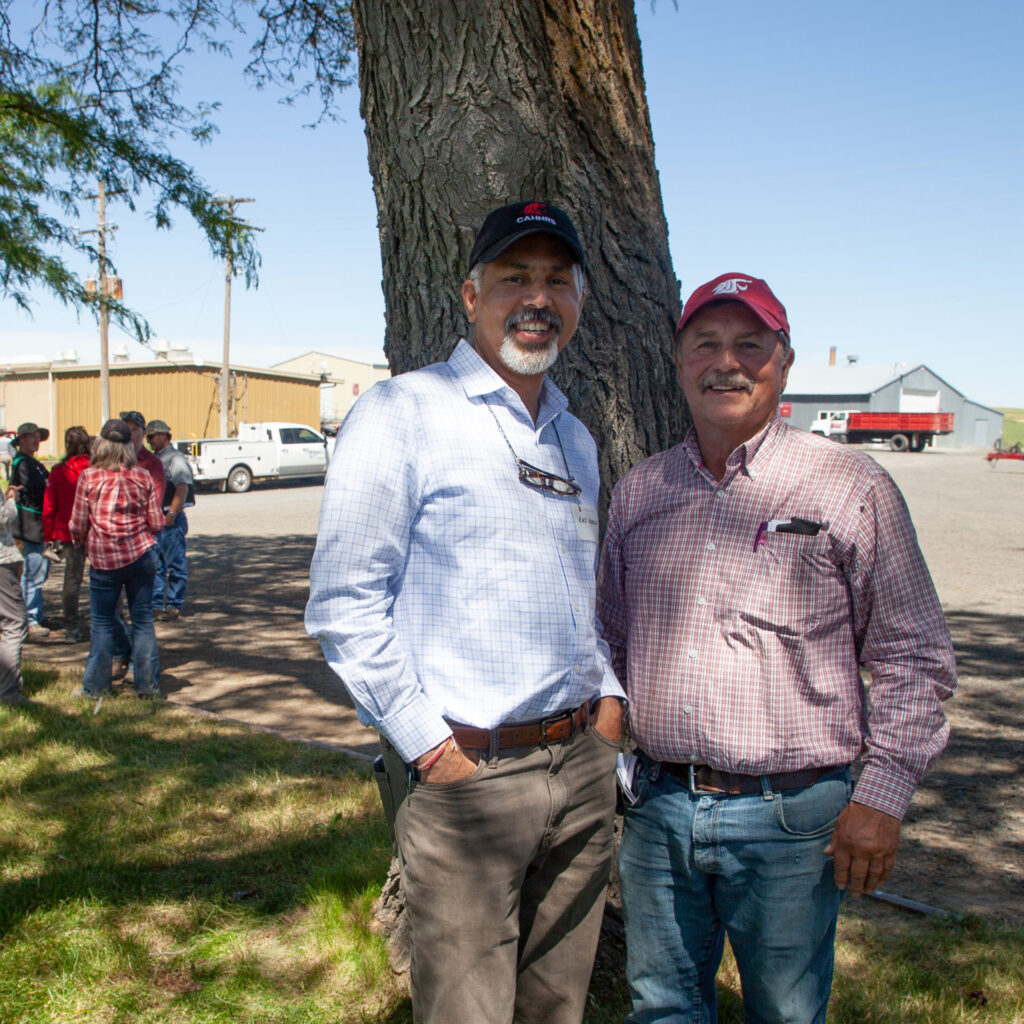 Two men stand together under a tree, smiling at the camera. There are buildings, trucks, and several other people in the background.