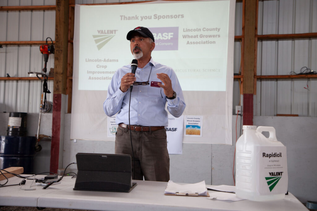 Dean Raj Khosla gestures while speaking into a microphone and standing behind a table with an iPad on it. On the table is a clipboard with papers on it, and a jug with a label that reads Rapidicil. On the projector screen behind him, text reads Thank you sponsors: Valent, BASF, Lincoln County Wheat Growers Association, Lincoln-Adams Crop Improvement Association.