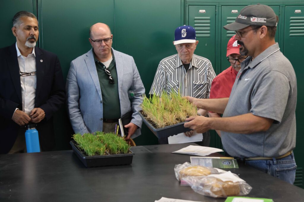 Five people stand in front of lockers and near a table. On the table are small grass plants in a tray. One of the people in the group holds up another tray of grass plants.