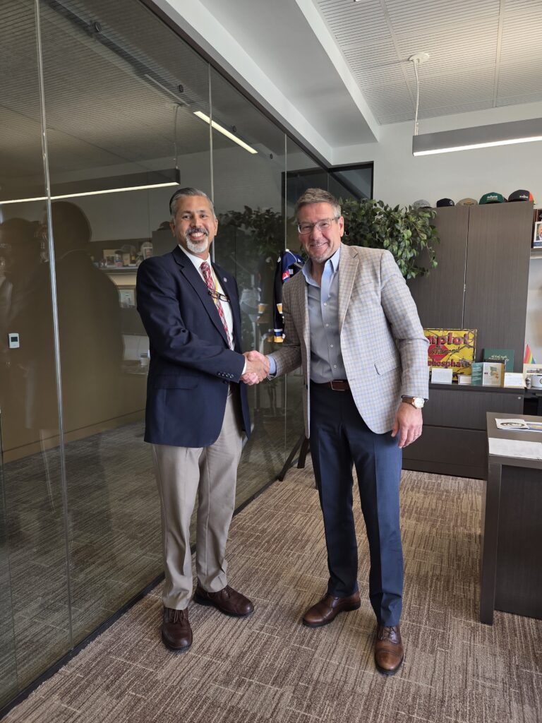 Two men shake hands in an office while smiling at the camera.