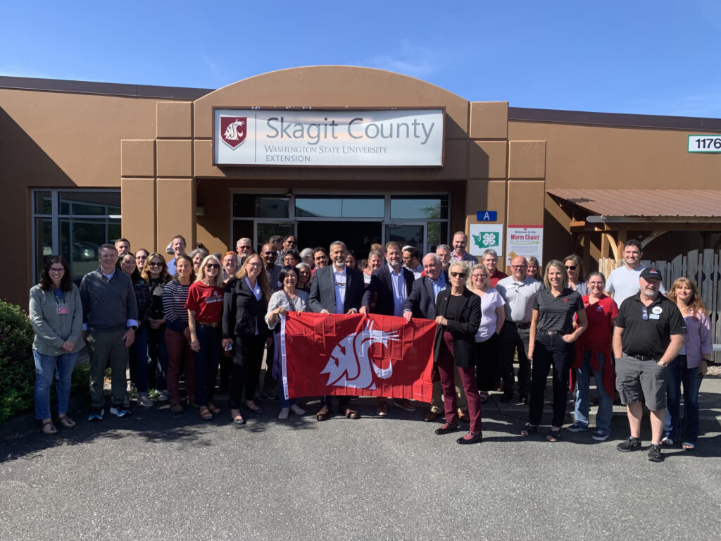 A large group of people stands in front of the WSU Skagit County Extension office. Some are holding a large WSU flag with the cougar head logo on it.