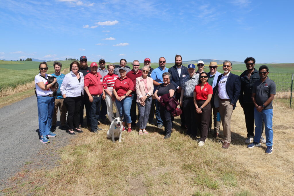 A group of 22 people stands together on a gravel driveway and grassy area near green fields.