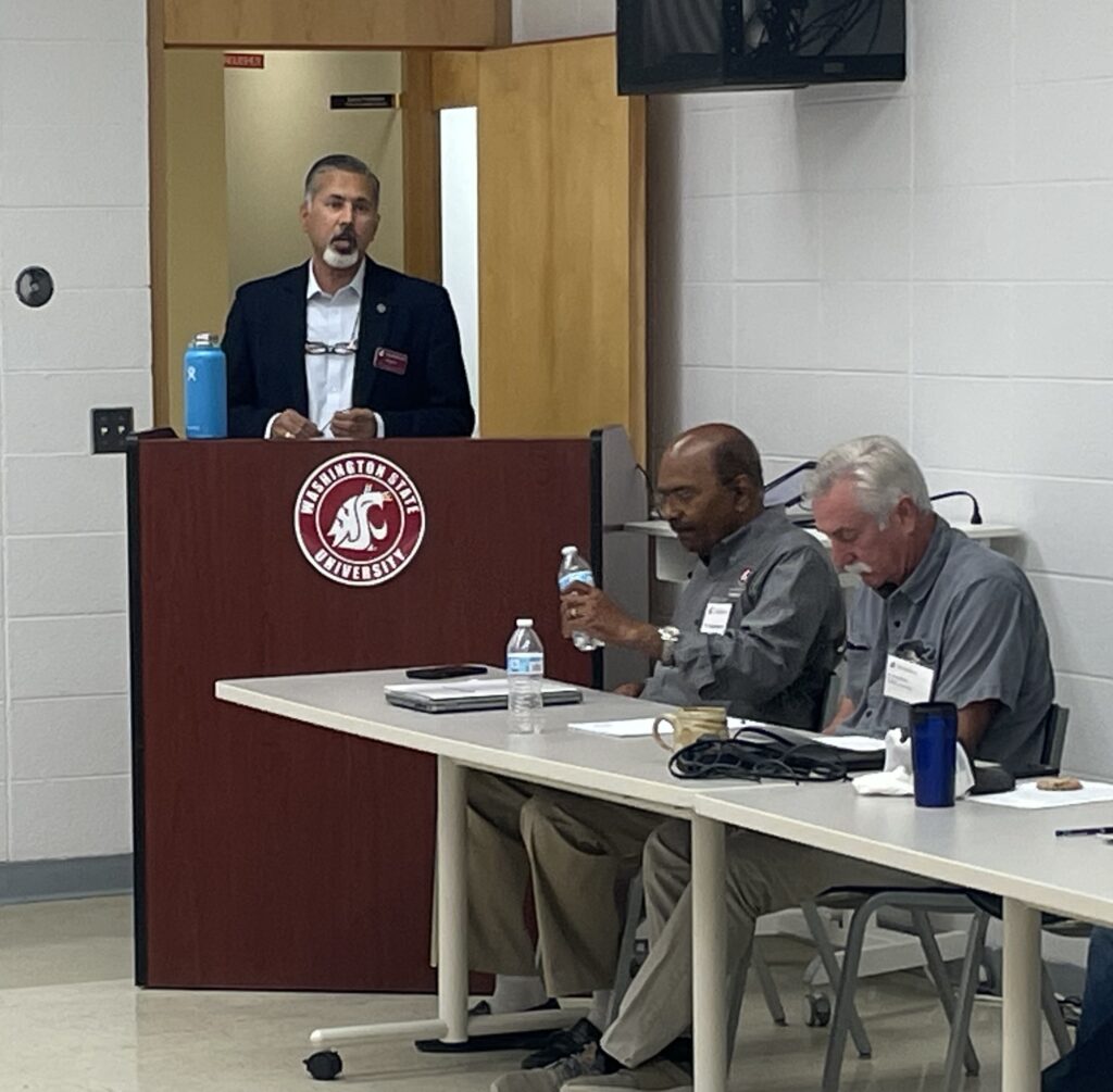 Raj Khosla stands at a podium with the WSU logo on it. Two people are seated at a table nearby.
