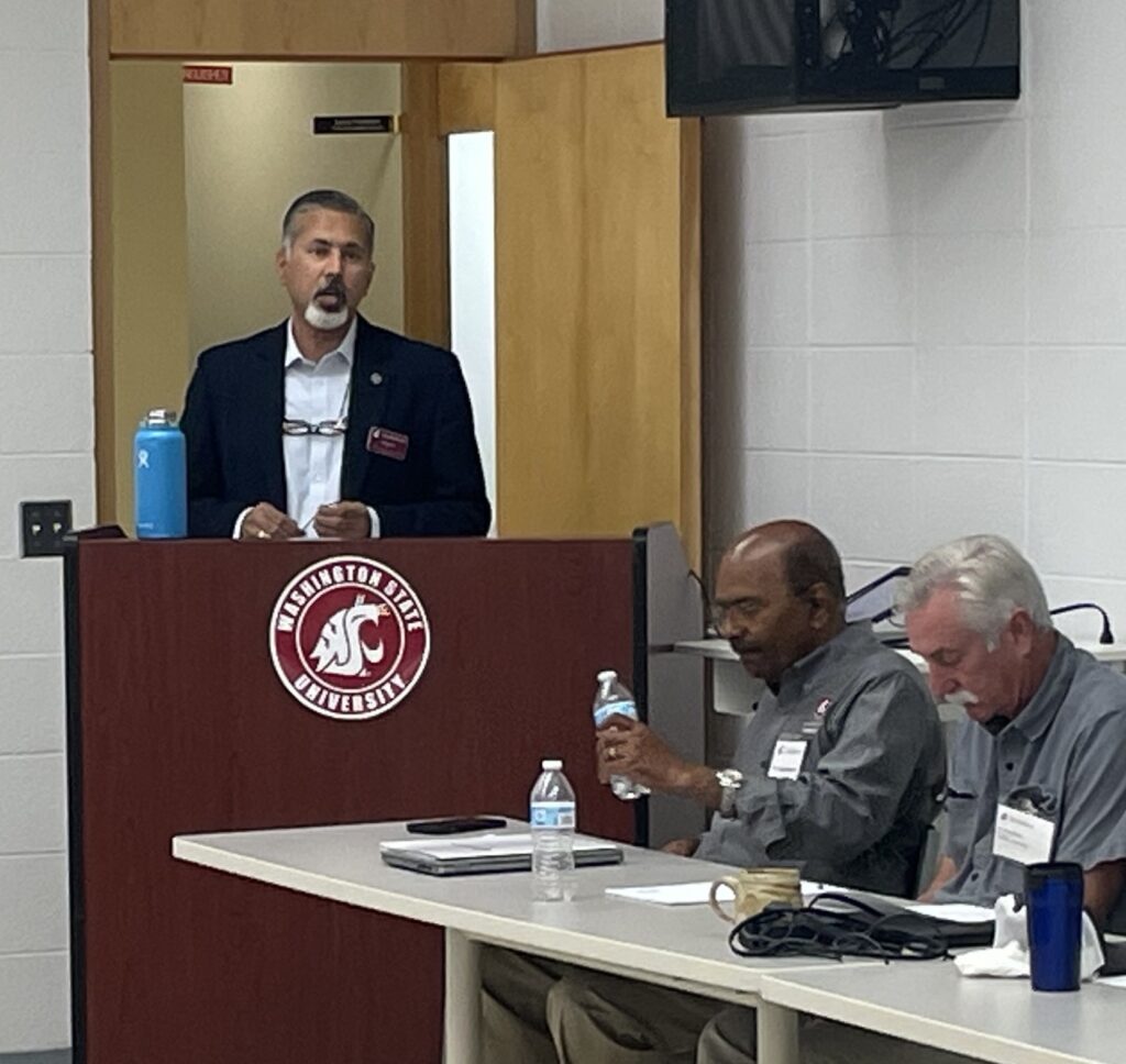 Raj Khosla stands at a podium with the WSU logo on it. Two people are seated at a table nearby.