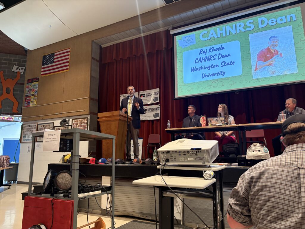 Raj Khosla stands at a podium on a stage, next to a projector screen that reads "CAHNRS Dean Raj Khosla CAHNRS Dean Washington State University." The screen also features a photo of Dean Khosla. Three people sit at a table near the podium on stage. In the foreground is a projector machine, a cart with equipment on it, and the back of a person.