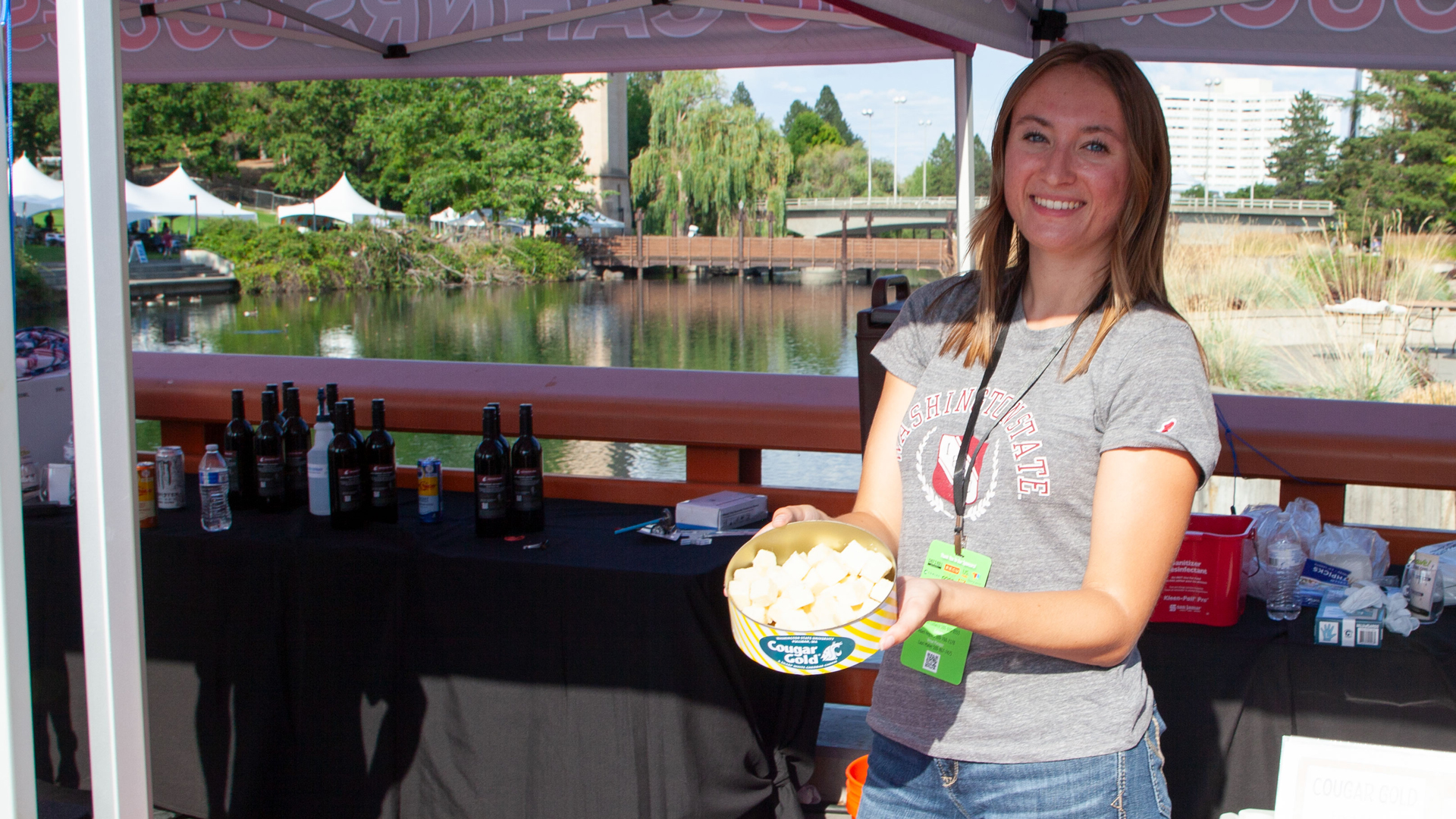 A person holds a can of Cougar Gold cheese samples while standing under a CAHNRS themed tent. On the table in front of her are another can of cheese samples in a bowl of ice and a can of beef stick samples in a bowl of ice.