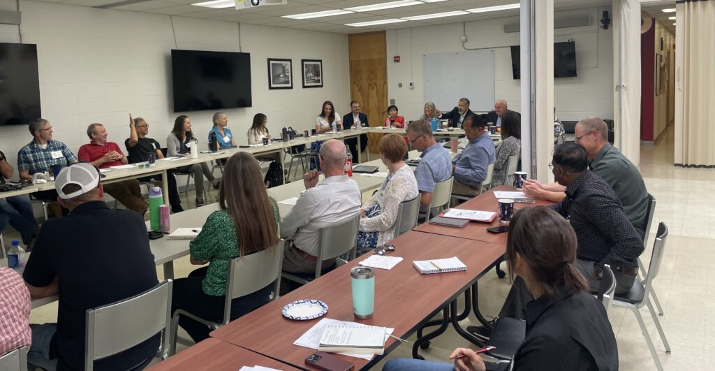 A couple dozen people are seated at tables in a large meeting room. Some have their backs to the camera while others are facing the camera.