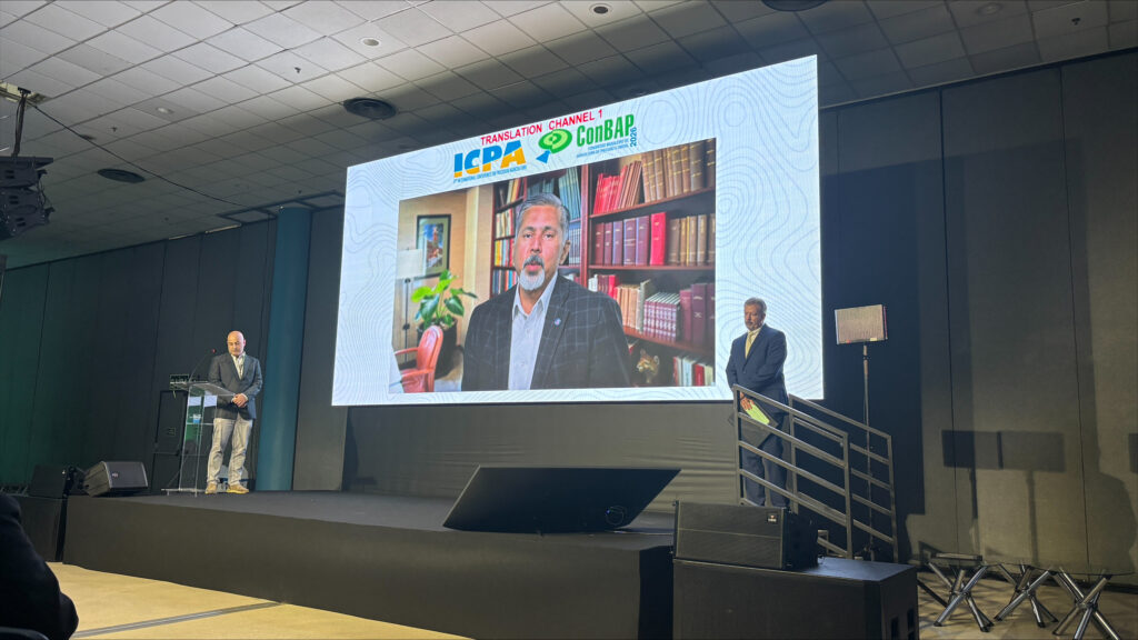 Raj Khosla appears on a projector screen above a stage during a conference. Two men stand on the stage, one on either side.