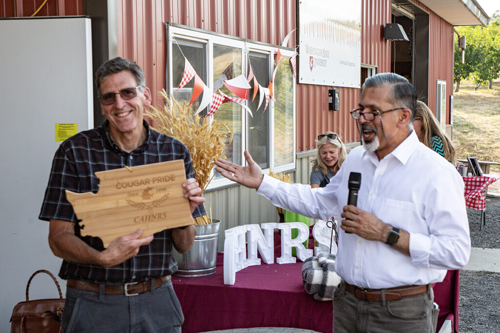 Raj Khosla speaks into a microphone while gesturing toward Scot Hulbert, who holds a WSU CAHNRS-themed cutting board shaped in the state of Washington. A red, metal building is behind them.