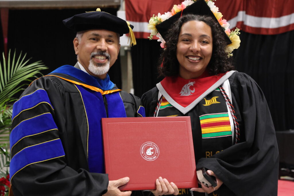 Dean Khosla in graduation regalia standing next to a student in cap and gown. Both hold a diploma folder.
